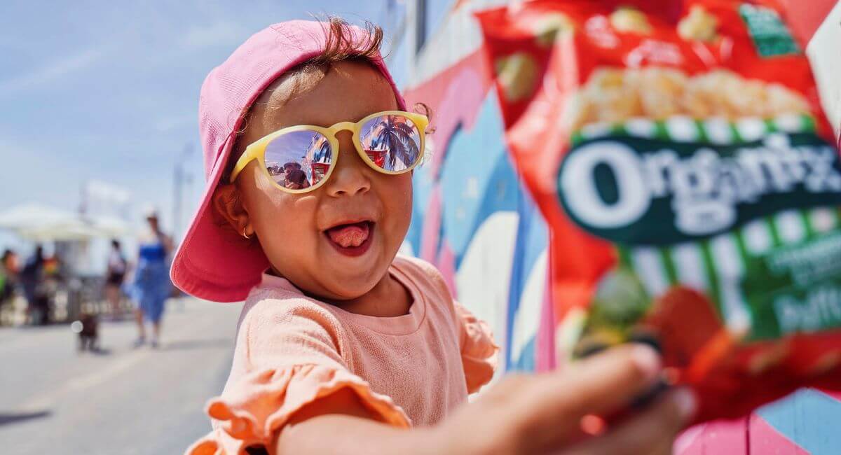 Happy child at the beach wearing a pink sunhat and sunglasses holding a bag of Organix puffcorn